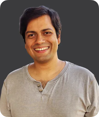 A photograph capturing an individual smiling and making direct eye contact with the camera. The person is wearing a grey t-shirt, and the image shows them from the shoulders up, with the smile prominently displayed. The background is neutral, allowing the person to stand out in the frame. The image conveys a warm and friendly expression.